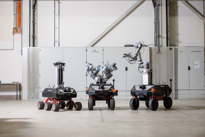 Three mobile robots stand in a factory hall. On the left is the six-wheeled sensor robot. In the middle is a mobile manipulator with two manipulators. On the right is a mobile manipulator with one manipulator attached to the mobile platform via a linear axis.