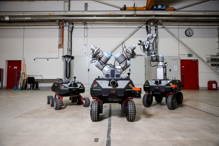 Three mobile robots stand in a factory hall. On the left is the six-wheeled sensor robot. In the middle is a mobile manipulator with two manipulators. On the right is a mobile manipulator with one manipulator attached to the mobile platform via a linear axis.