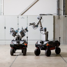 Two mobile manipulators are standing in a factory hall. The left mobile manipulator has two manipulators. The right mobile manipulator has one manipulator attached to the mobile platform via a linear axis.